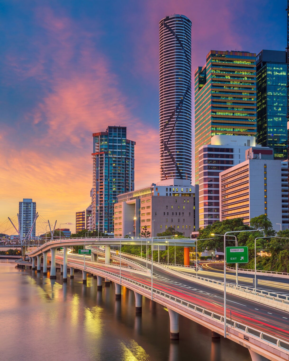 Brisbane skyline at sunset with illuminated buildings and a riverside highway.