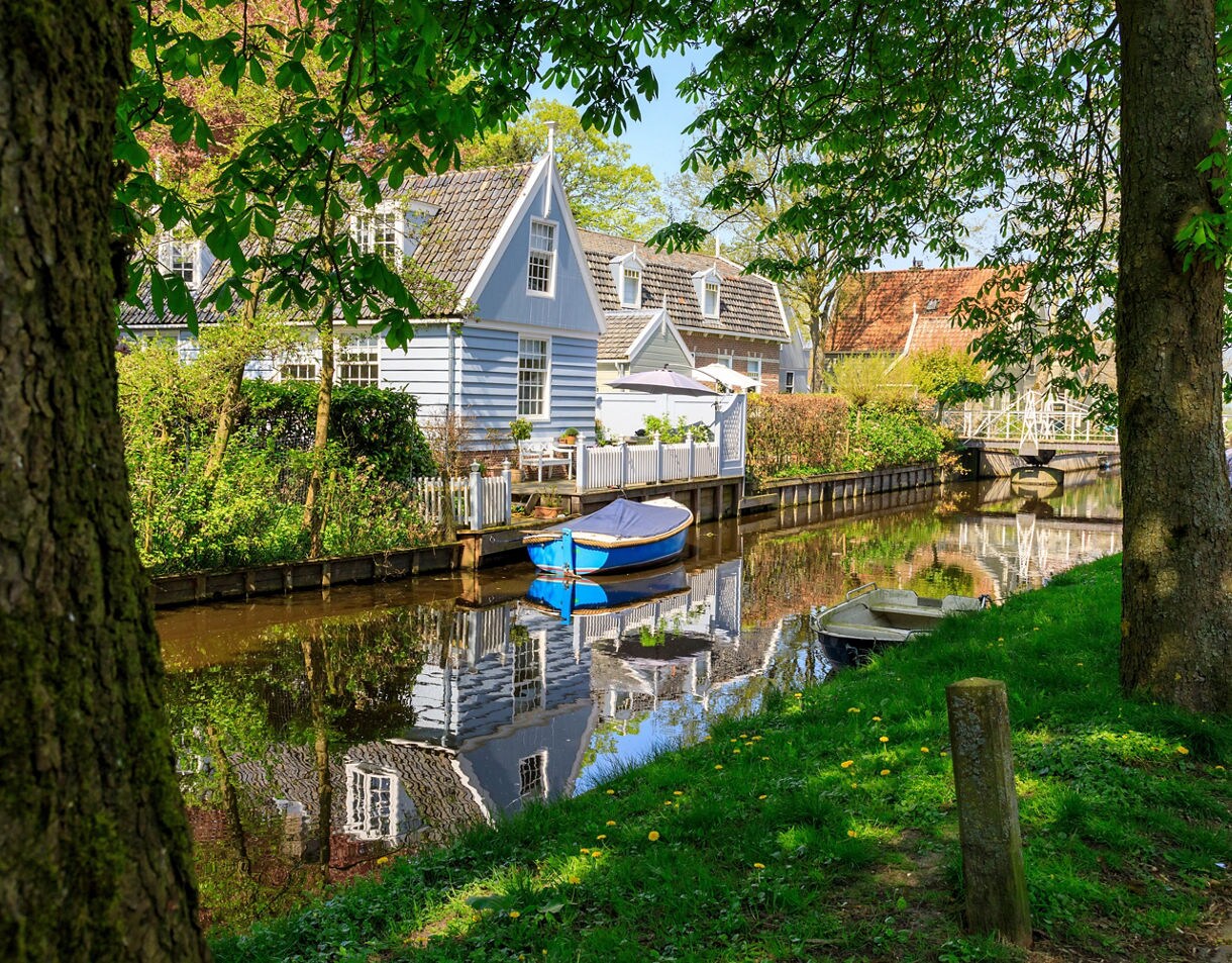 Small canal with boats and pastel wooden houses in Broek in Waterland surrounded by greenery.