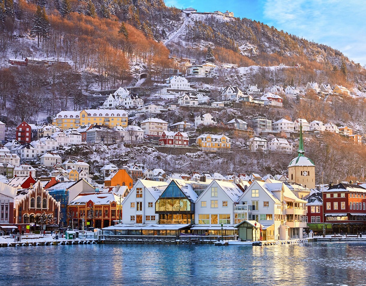 Snowy hillside in Bergen with colorful houses, church steeples and winter trees reflecting in the harbor.
