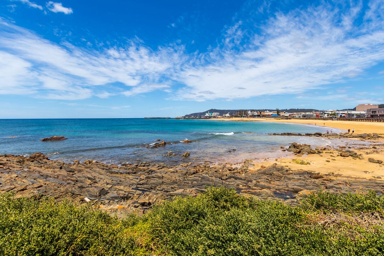 Rocky shoreline with golden sand and clear blue water near a small coastal town under a bright sky.