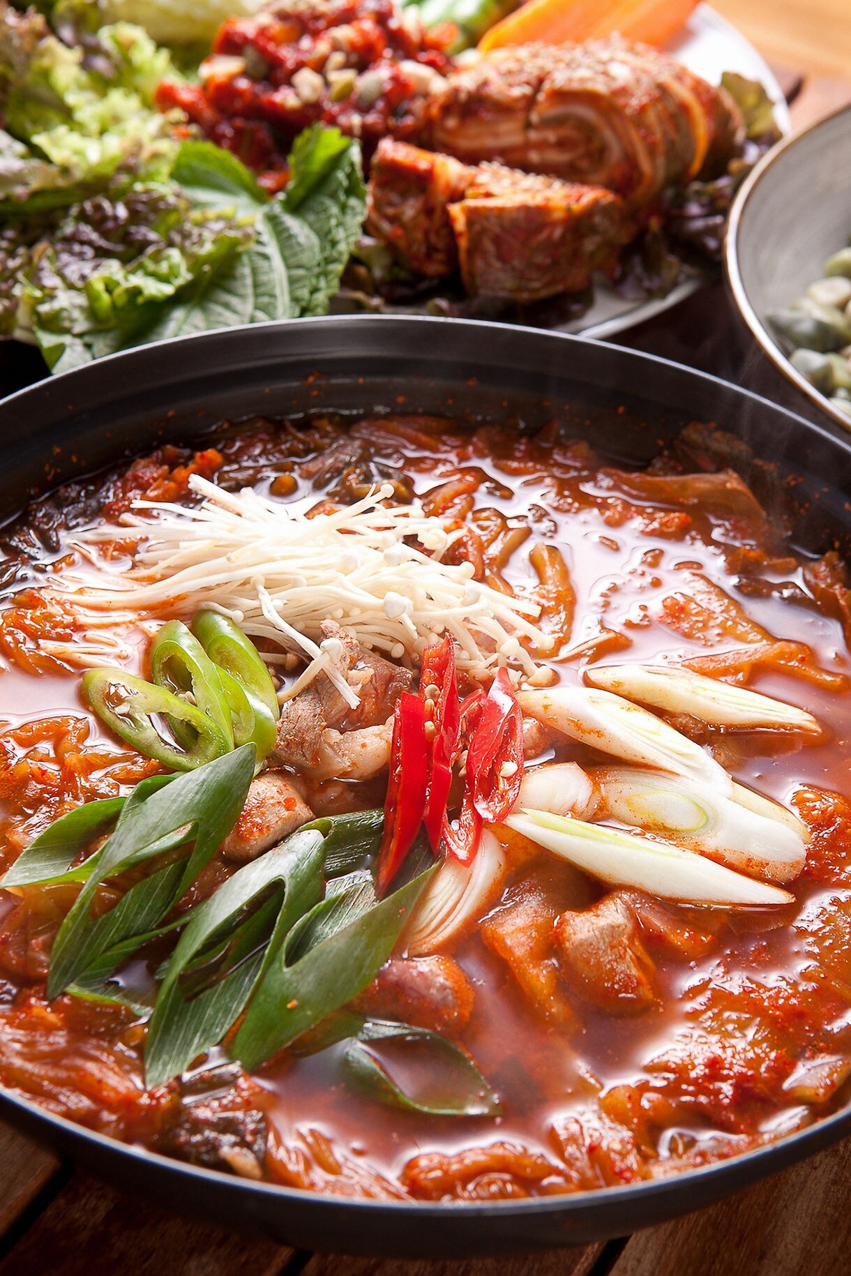 A cast iron pot containing a red spicy Korean stew with tofu slices, vegetables, and broth, accompanied by grilled meat and lettuce leaves on a dining table.