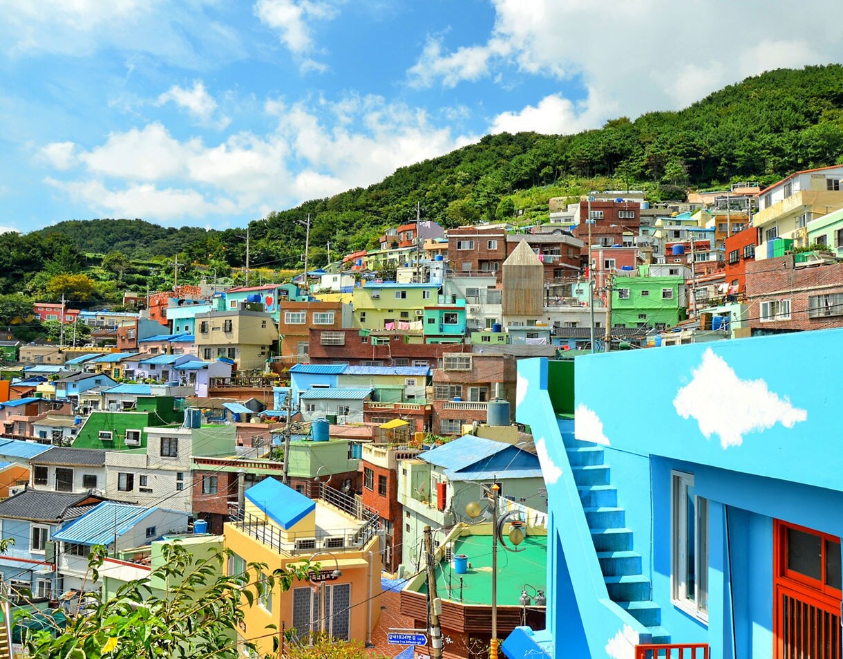 Hillside neighborhood filled with colorful houses and painted buildings, set against green forested hills and a bright blue sky.