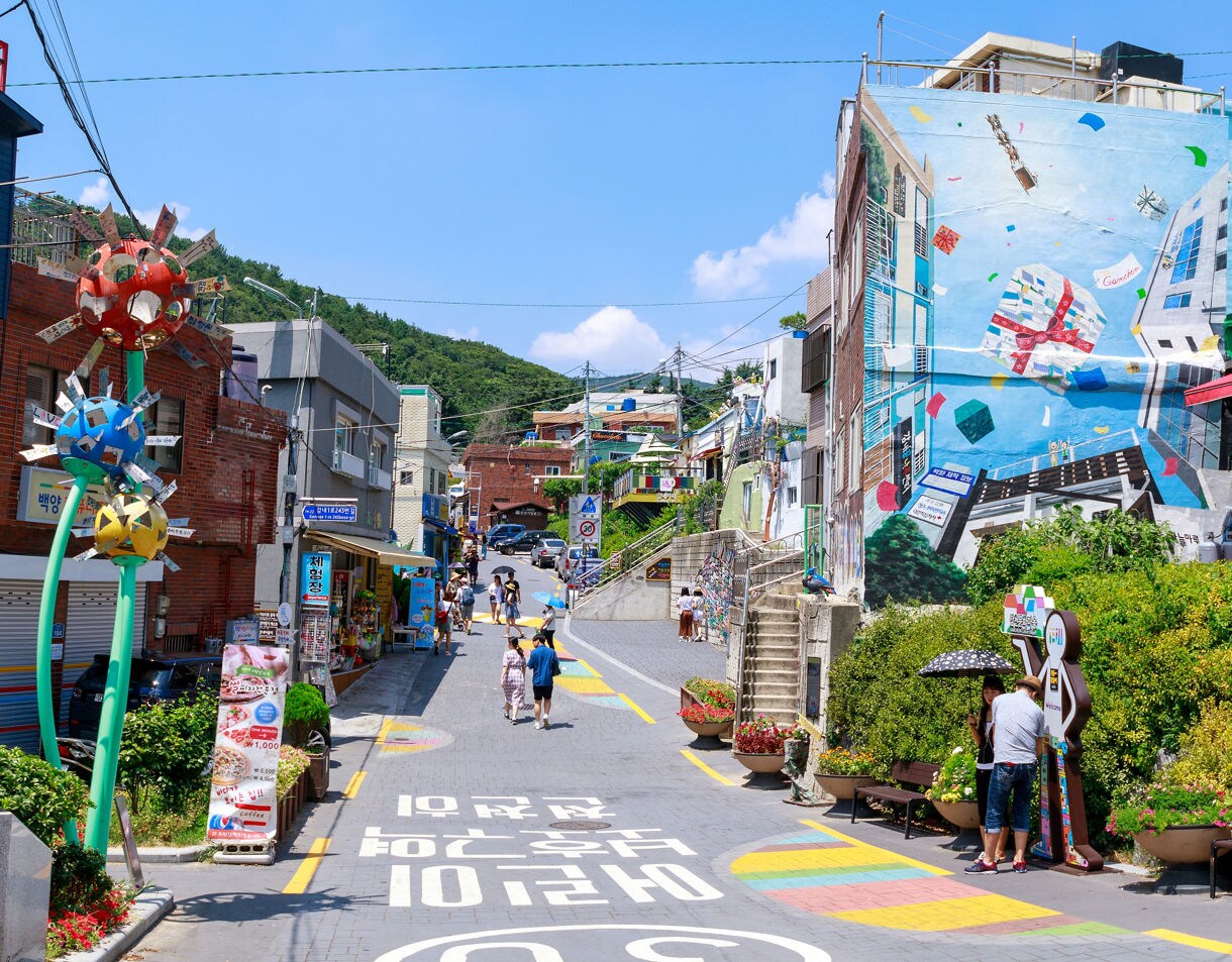 Street in Gamcheon Culture Village, Busan, lined with colorful buildings, wall murals and whimsical street art under a bright blue sky.