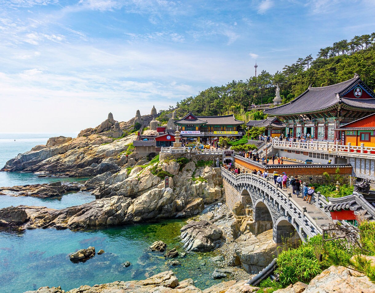 Colorful temple buildings perched on rocky cliffs beside the ocean, with a stone bridge leading to Haedong Yonggungsa in Busan.