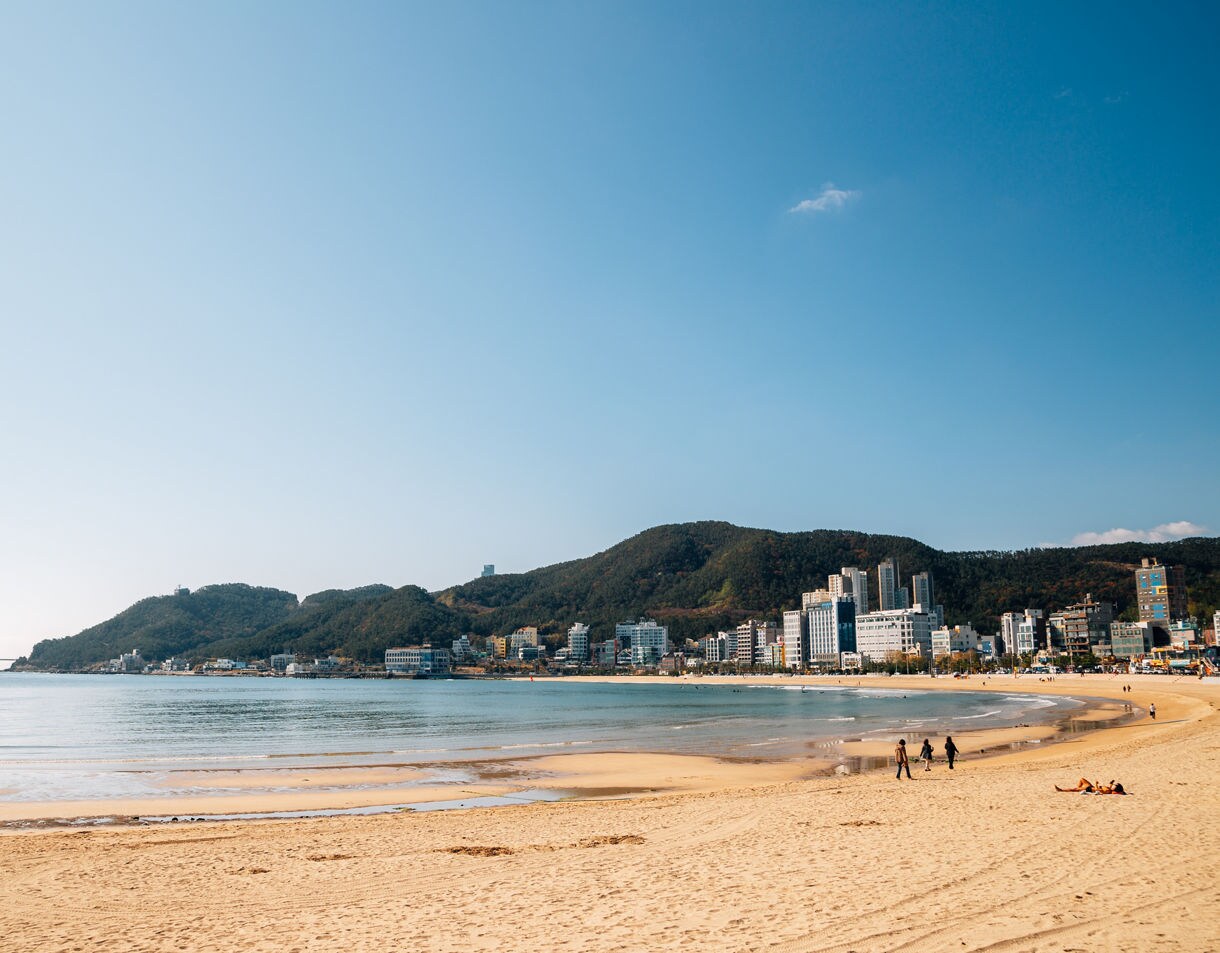 Wide sandy beach curving along the shoreline with calm waves, city buildings and forested hills in the background.
