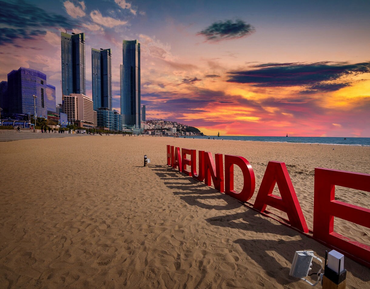 Large red “HAEUNDAE” letters on a sandy beach at sunset with tall buildings and colorful sky in the background.