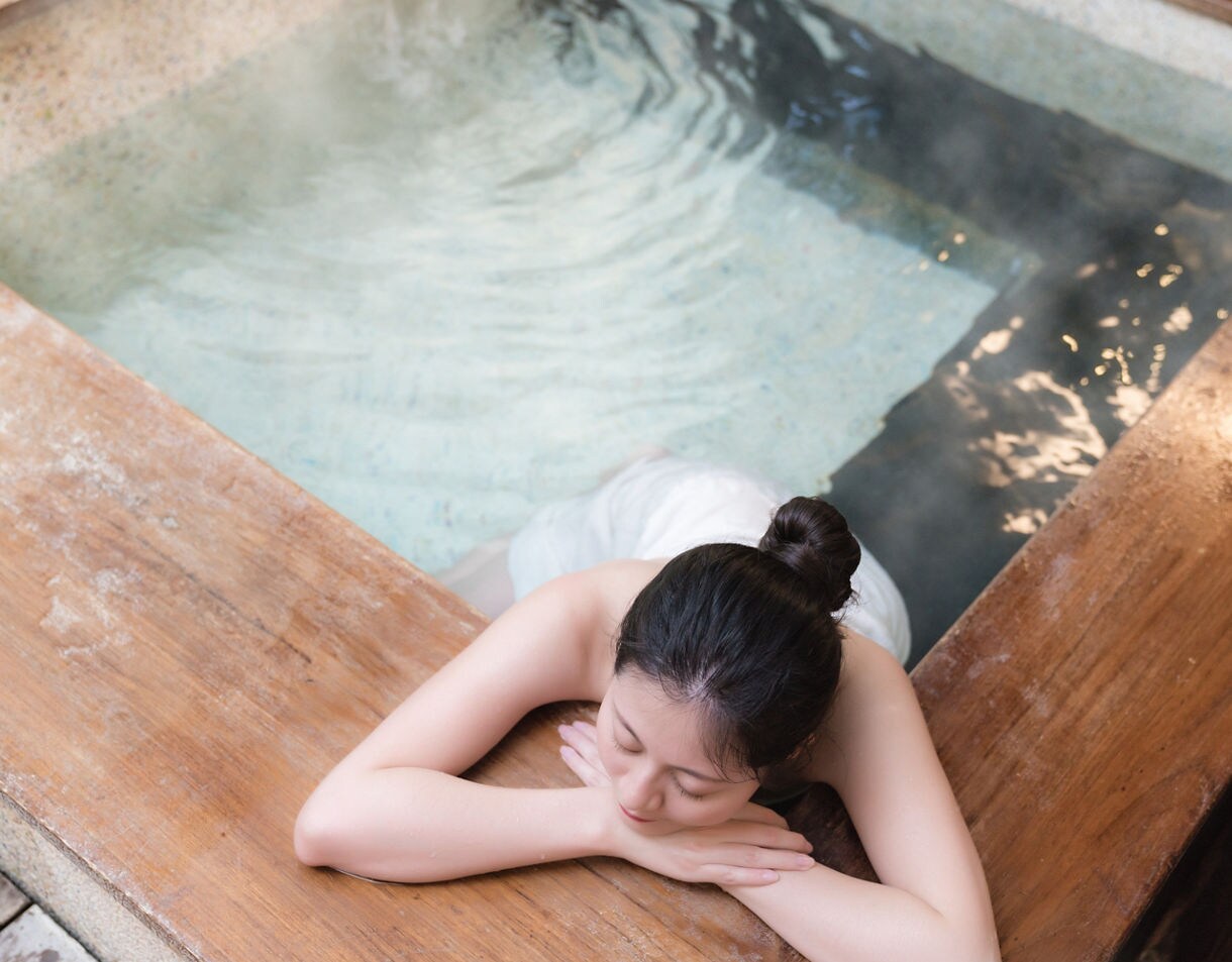 Woman relaxing at the edge of a warm, steamy spa bath at Spa Land in Busan, wrapped in a towel with eyes closed.