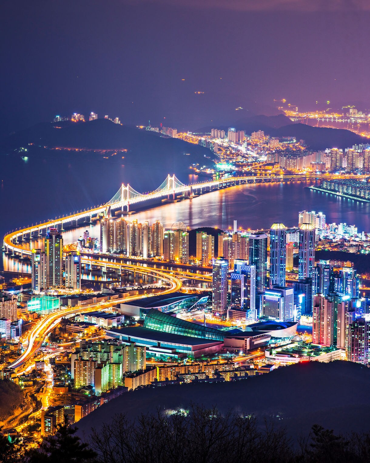 Aerial night view of Busan with illuminated skyscrapers, highways and the glowing Gwangandaegyo Bridge stretching over the water.