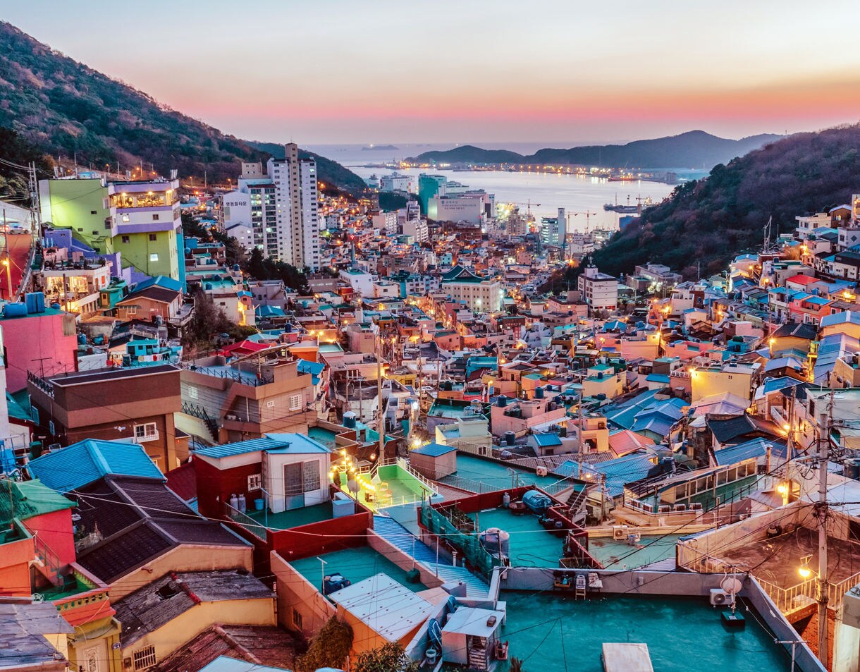 Colorful rooftops and winding streets of Busan at dusk, with city lights beginning to shine and the harbor in the distance.
