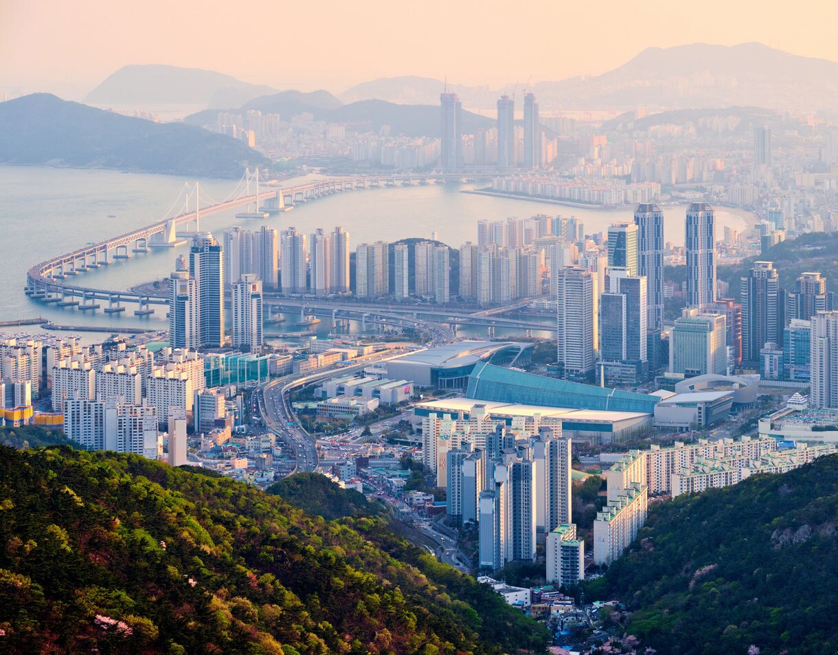 Panoramic cityscape of Busan seen from X the Sky observatory, featuring tall skyscrapers, Gwangan Bridge and surrounding mountains at sunset.