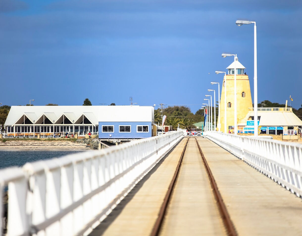 View down the long white Busselton Jetty with rail tracks, leading toward coastal buildings and a lighthouse under a bright blue sky.