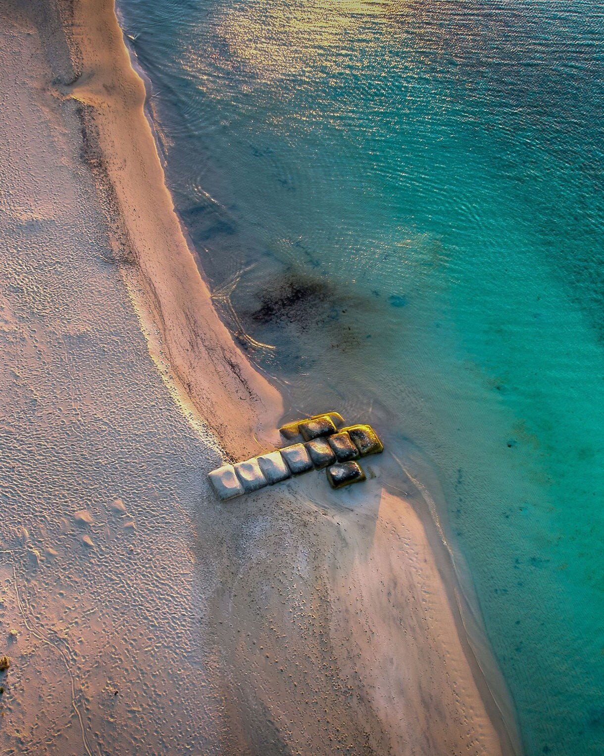 Aerial view of a sandy coastline with scattered vegetation and clear turquoise water, where a small cluster of concrete blocks sits near the shoreline.