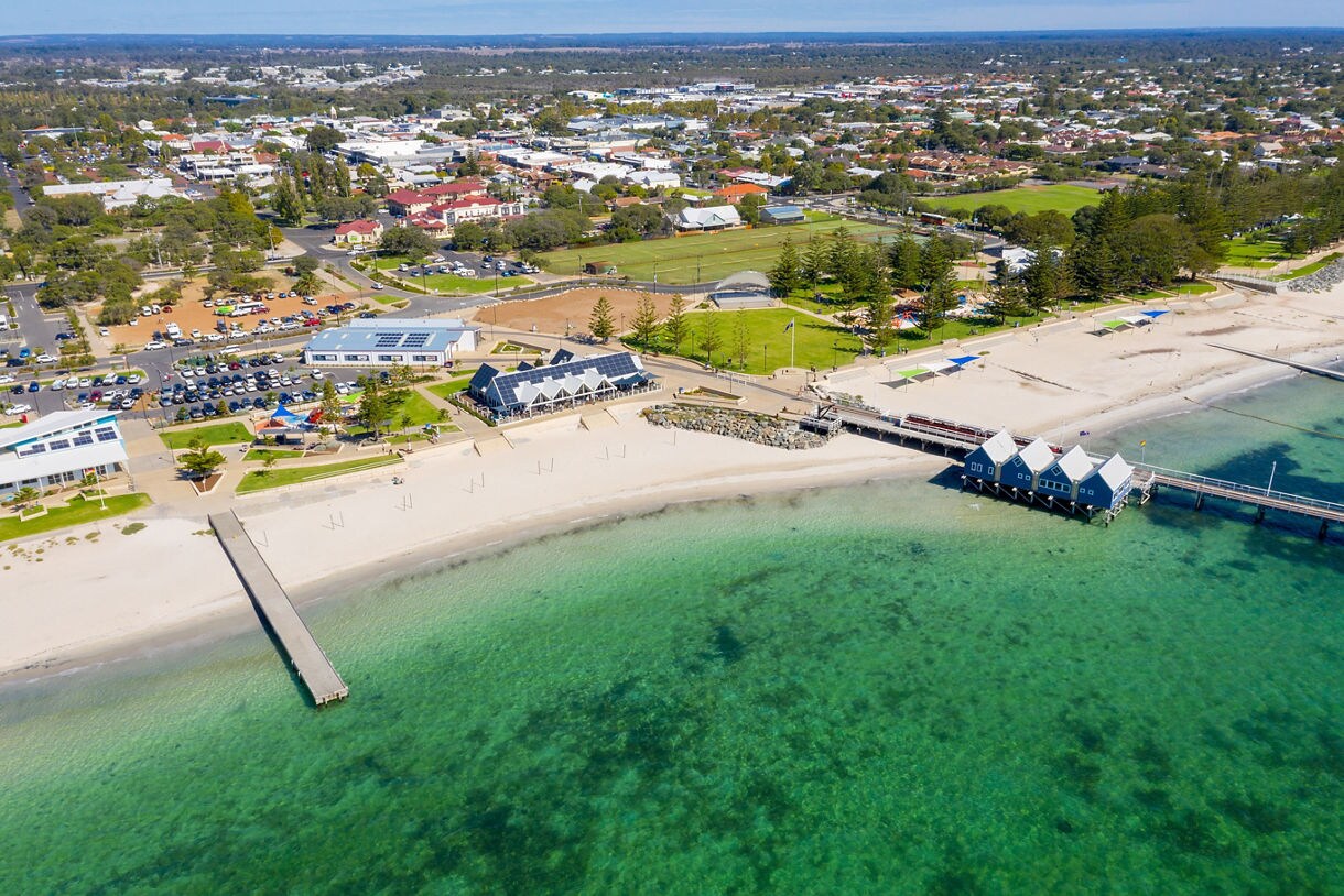 Aerial view of Busselton Foreshore with white sand beaches, clear green-blue water, grassy park areas and the long Busselton Jetty extending into the ocean.