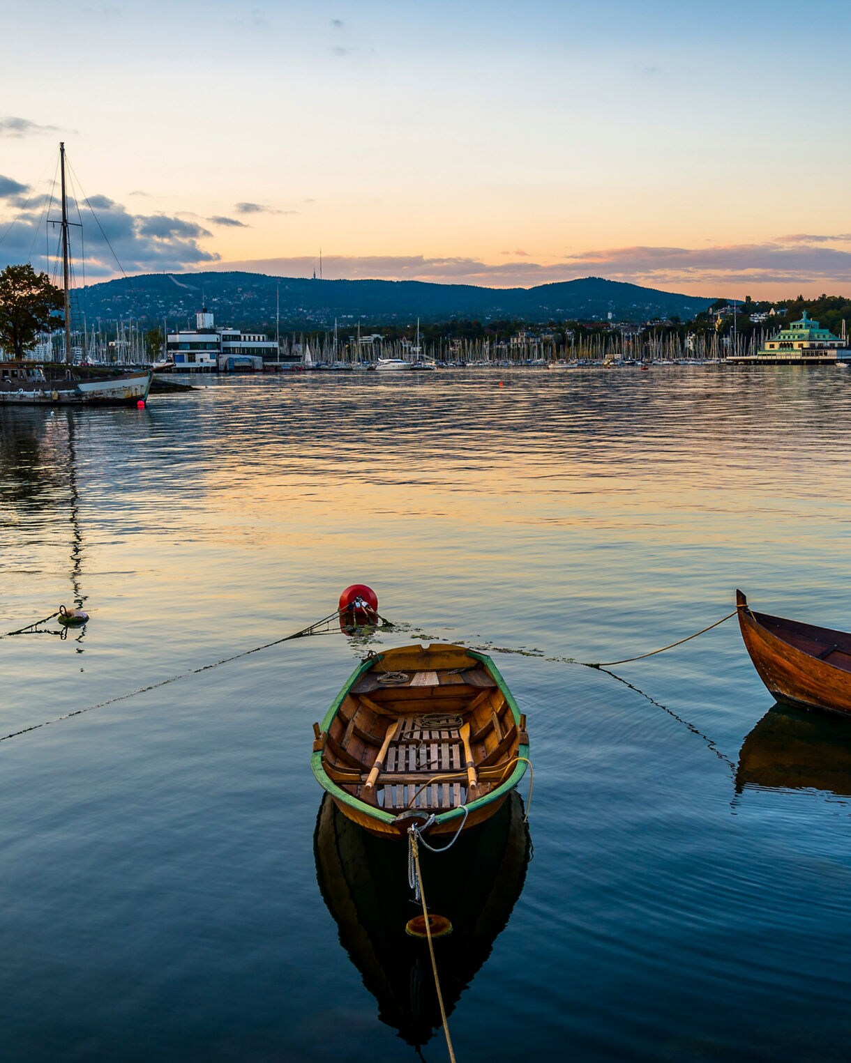 Tranquil view of wooden boats moored at Bygdøy Peninsula in Oslo, Norway, with calm water reflecting the sunset sky and distant hills.