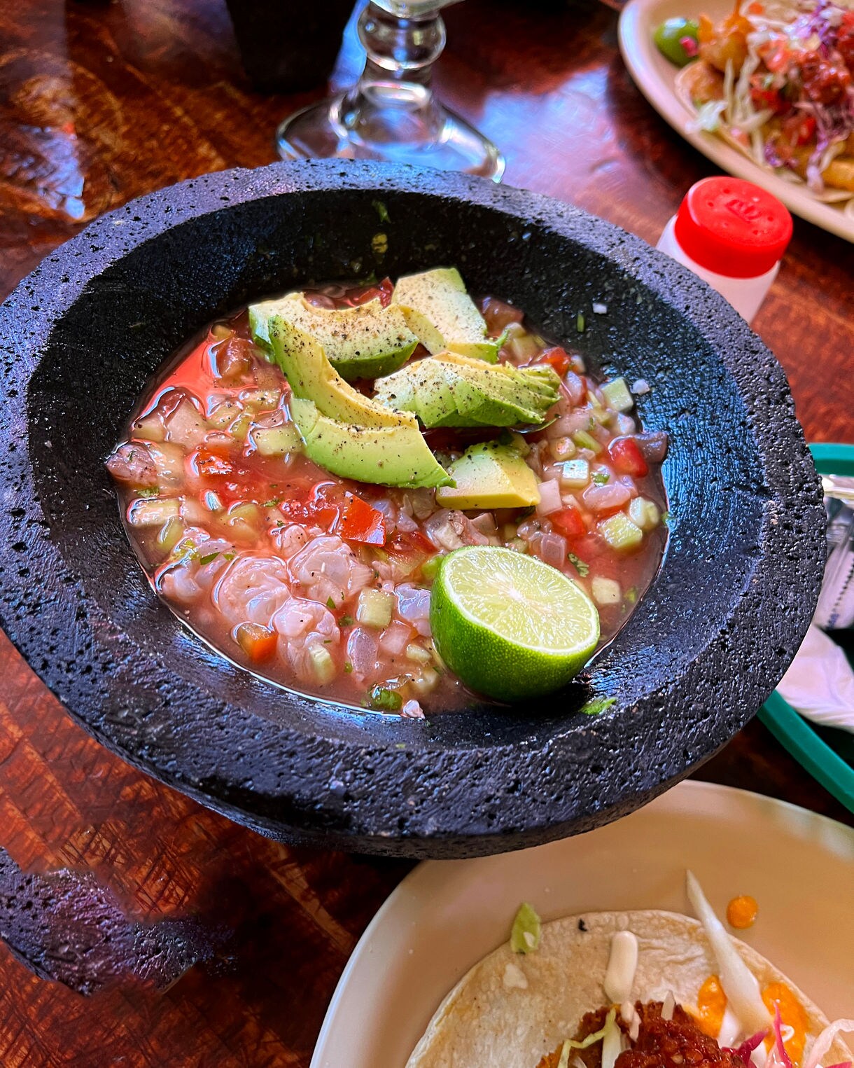 Bowl of shrimp ceviche with diced tomatoes, cucumbers, avocado slices and lime in a black stone dish on a wooden table.