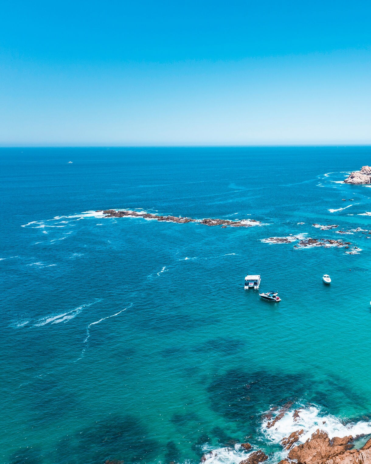 Aerial view of Chileno Bay in Cabo San Lucas with turquoise waters, anchored boats, sandy beach and rocky shoreline.