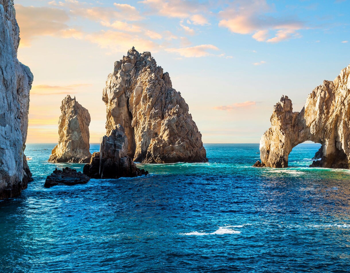 Dramatic view of El Arco at Cabo San Lucas during sunset, with rugged rock formations above deep blue waters and a glowing sky.