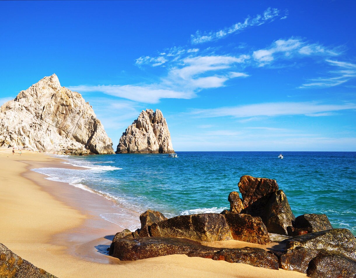 Wide sandy beach at Cabo San Lucas with rocky formations and turquoise waves under a bright blue sky.