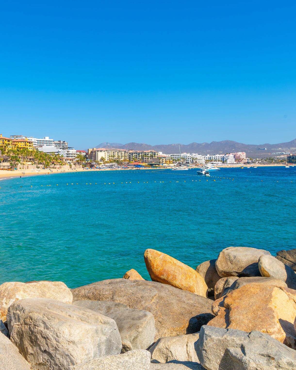 Medano Beach in Cabo San Lucas lined with resorts, sunbathers, umbrellas and gentle waves along the golden shoreline.