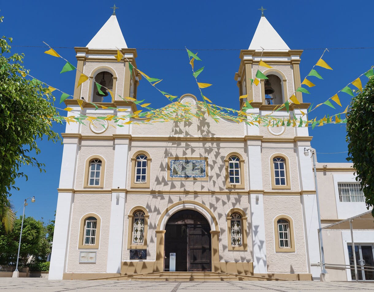 Front view of the Mission of San José del Cabo featuring twin bell towers, arched doorway and festive green and yellow banners strung across the plaza.