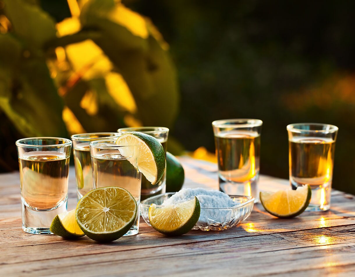 Glasses of golden tequila on a wooden table with lime wedges and a small dish of salt, lit by warm sunlight.