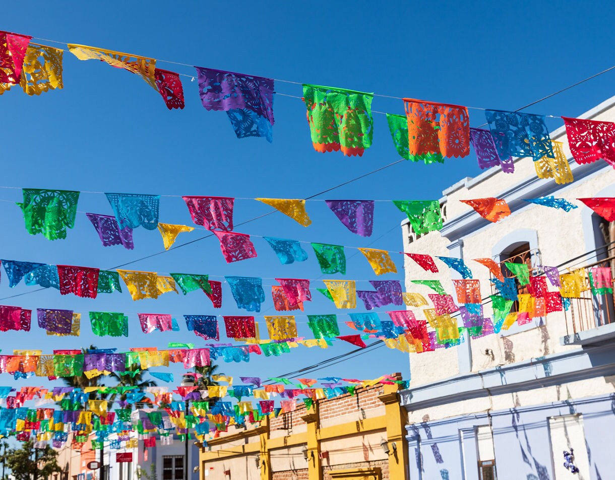 Rows of colorful papel picado banners strung across a street in Todos Santos with blue sky and colonial-style buildings in the background.