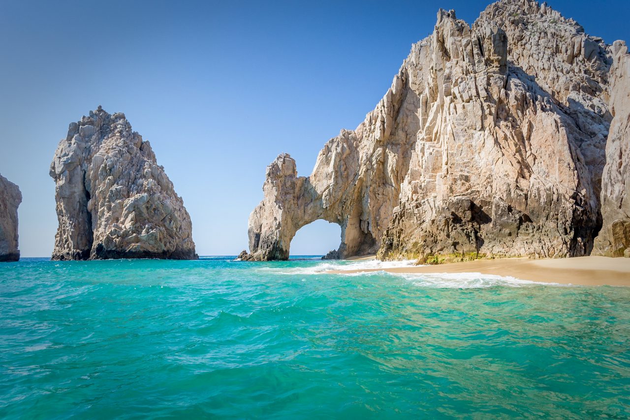 Natural rock arch formation at Cabo San Lucas rising above clear blue waters, with golden sand and rugged cliffs under a bright sky.