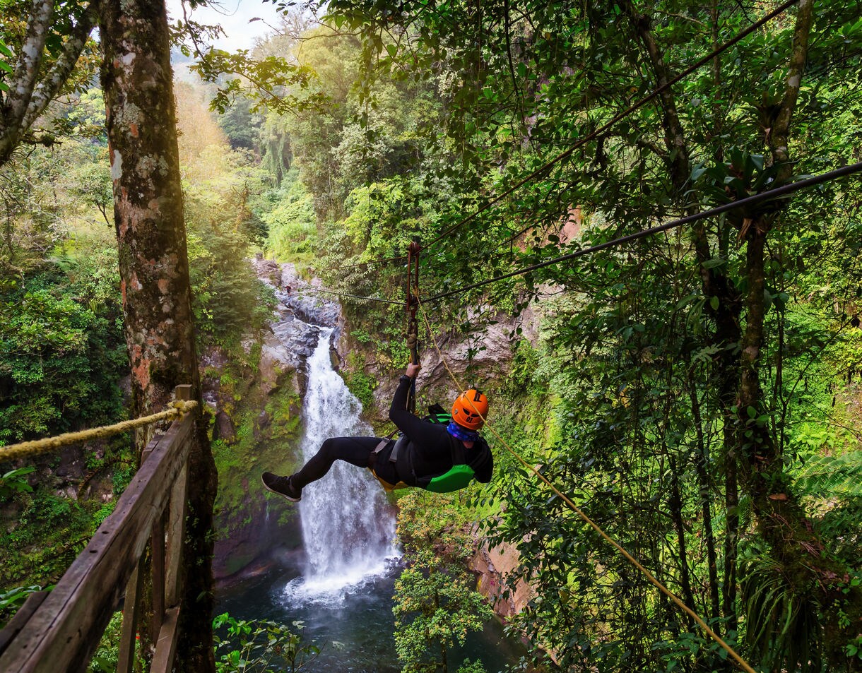 Person ziplining through lush green forest in Cabo with a tall waterfall cascading into a pool beneath.