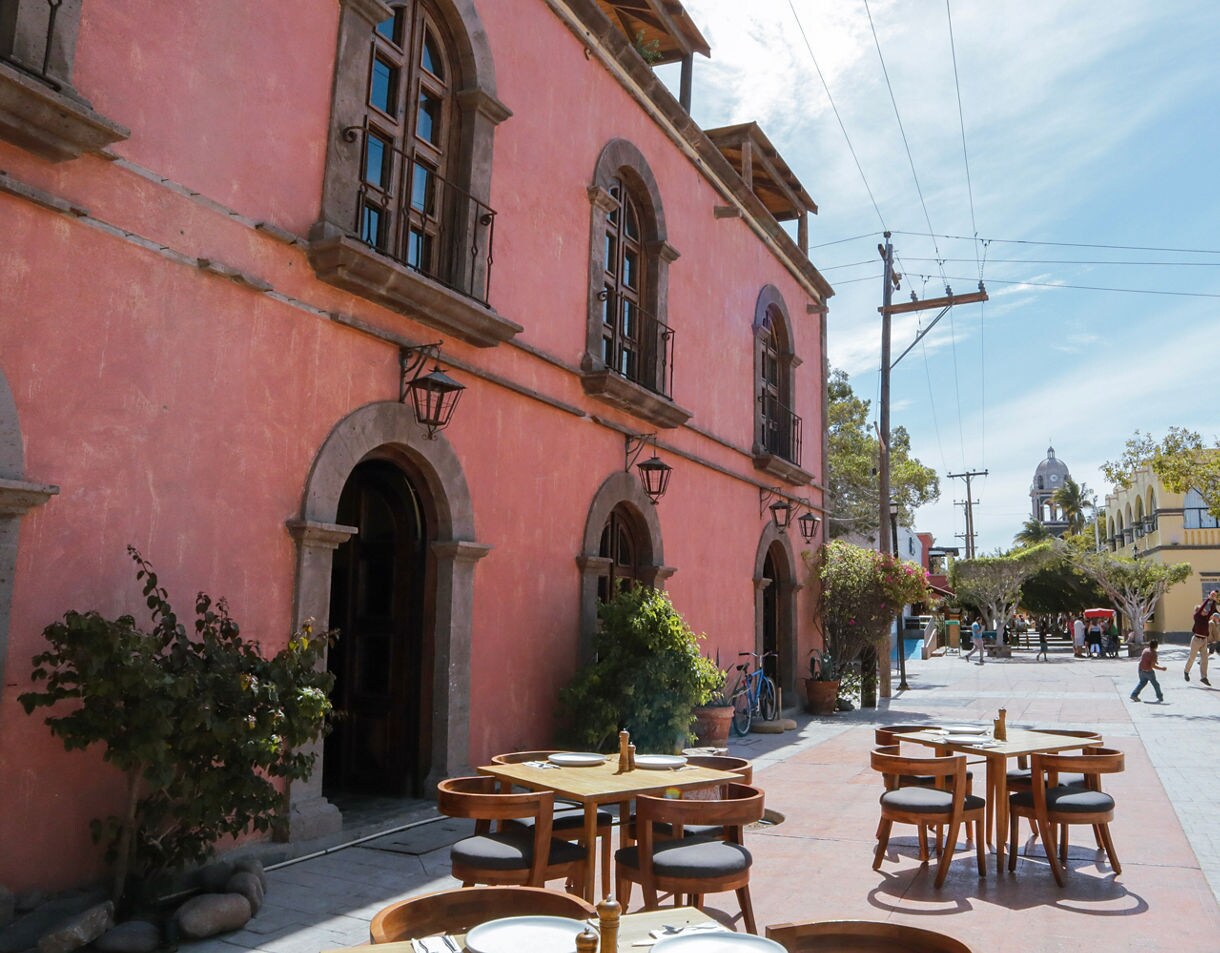 Outdoor café tables set along a pink colonial building in Baja’s old town, with people walking and playing in a sunlit plaza nearby.