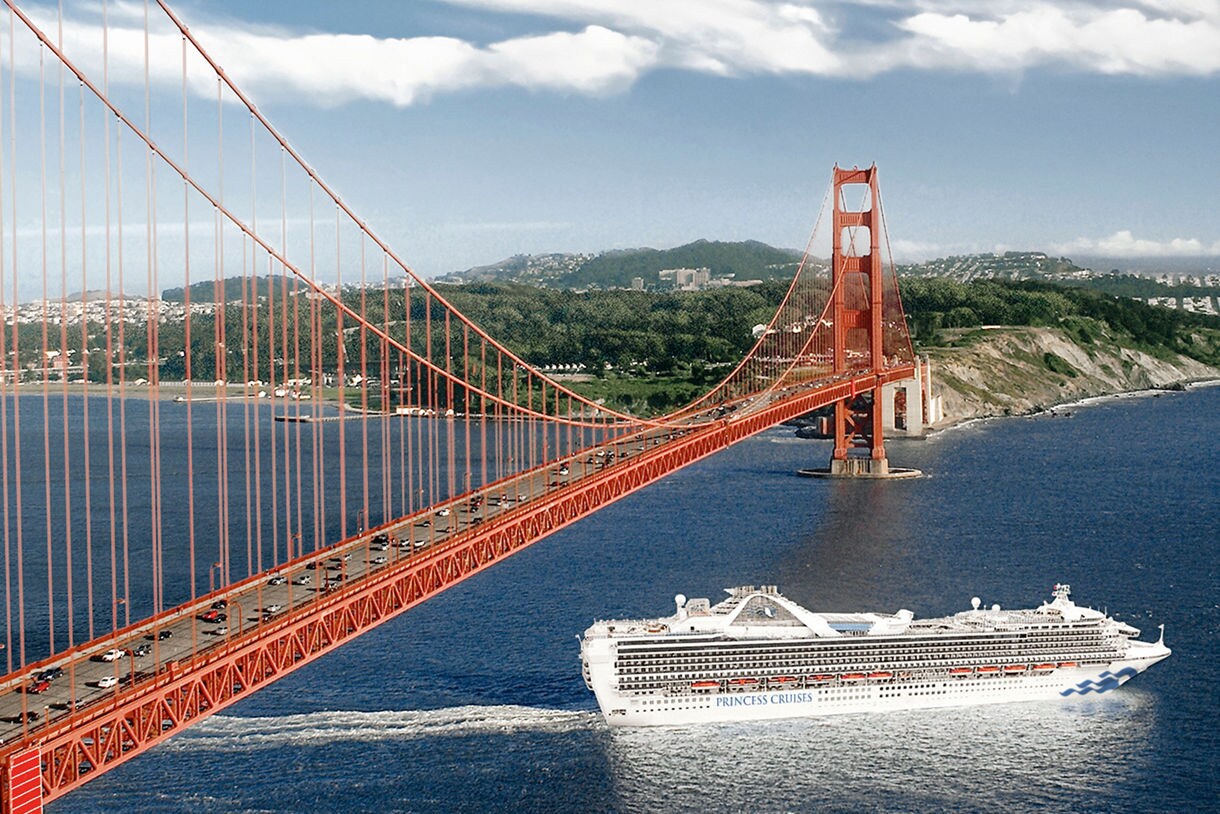 A Princess Cruises ship passes beneath the Golden Gate Bridge in San Francisco Bay, with green hills and residential areas visible in the background.