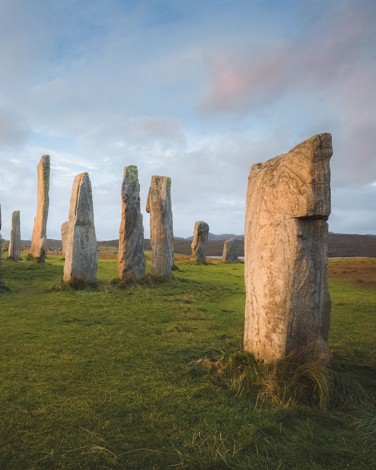 Wide view of the Callanish Standing Stones with tall weathered monoliths arranged in a circle on a grassy field during a pastel sunrise.