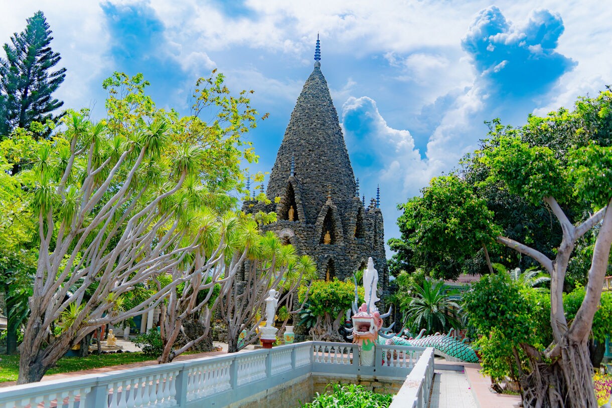  A view of the unique Tu Van Pagoda in Cam Ranh, Vietnam, which is a Buddhist temple complex notably featuring a large tower built from millions of coral and shell pieces. The pagoda is surrounded by lush tropical vegetation and a reflective pond or walkway in the foreground.