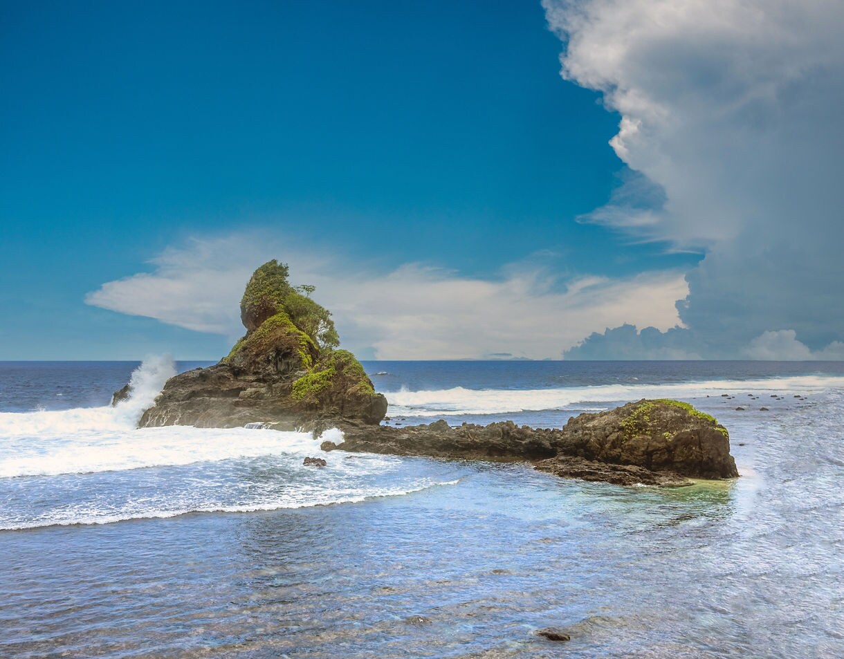 Rocky coastal formation covered in small patches of greenery, surrounded by blue ocean waves and a partly cloudy sky.