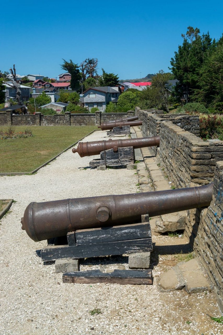 Row of rusted historic cannons mounted on wooden supports along a stone wall in an outdoor fort, with houses and trees visible in the background.