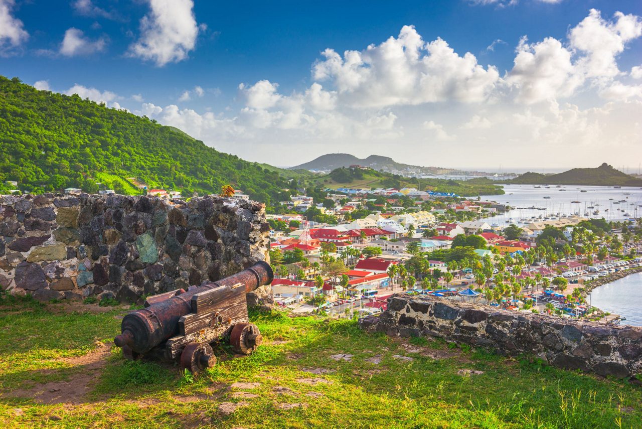 Historic stone fort ruins with old cannons in foreground overlooking tropical bay with colorful houses, green hills, and boats in harbor below