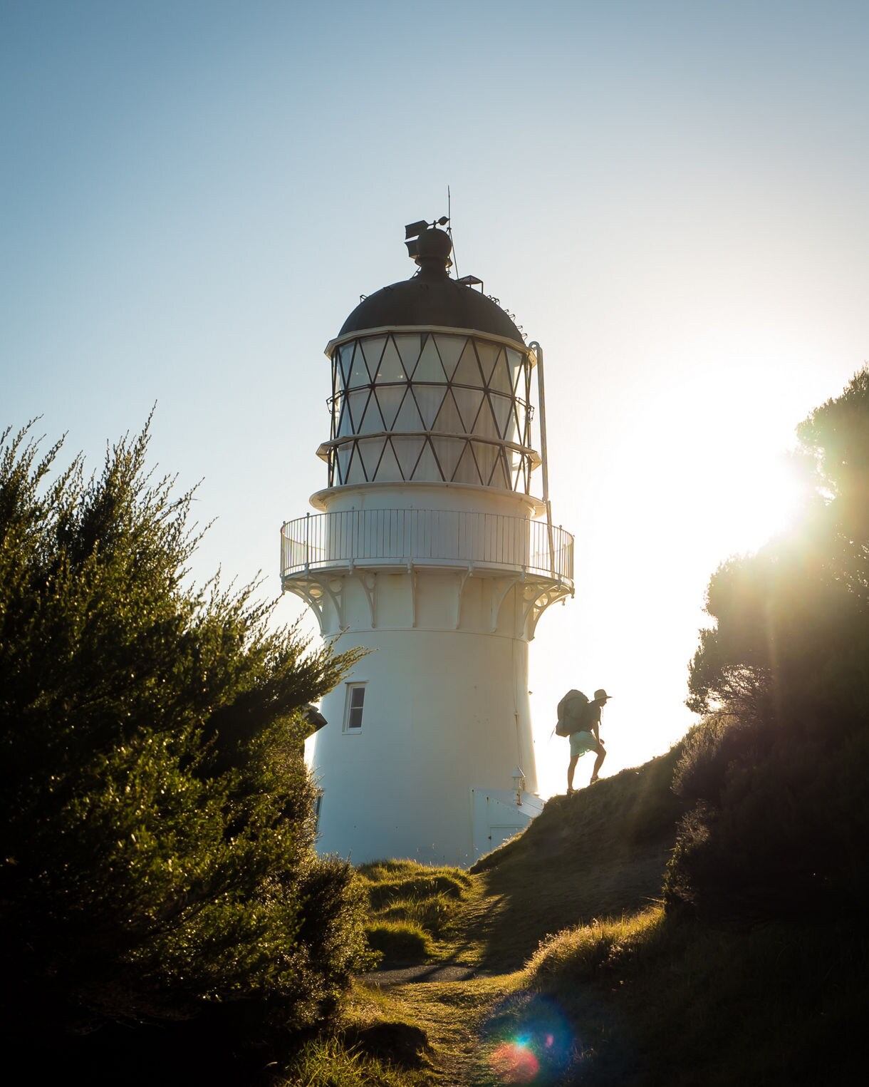 Silhouette of a hiker climbing a grassy path toward the white Cape Brett Lighthouse, backlit by the warm evening sun.