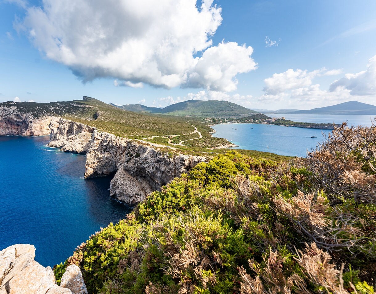 High limestone cliffs rising above deep blue sea with green scrubland and distant hills under bright clouds.