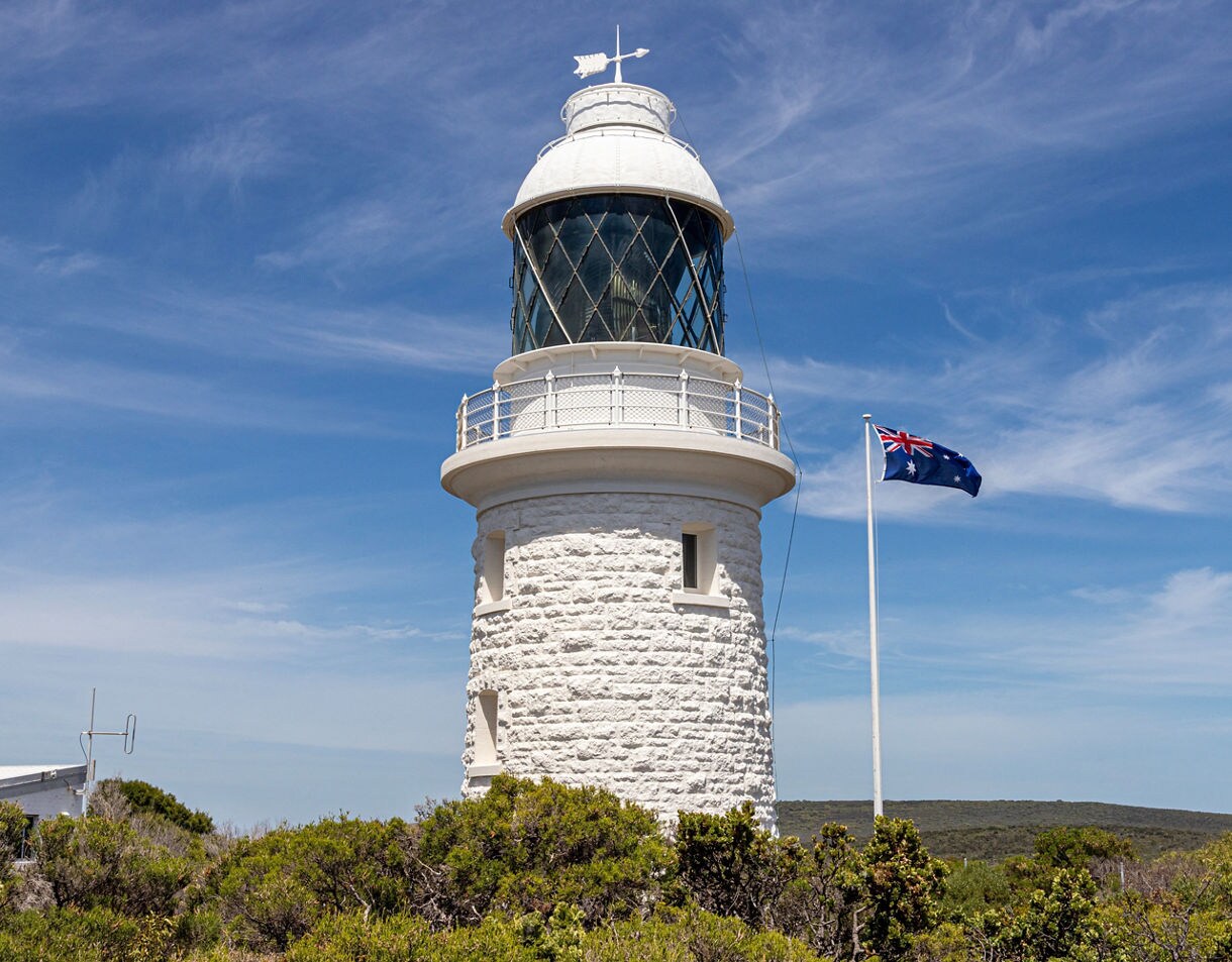 White stone lighthouse at Cape Naturaliste with an Australian flag flying nearby, surrounded by low coastal vegetation under a bright blue sky.