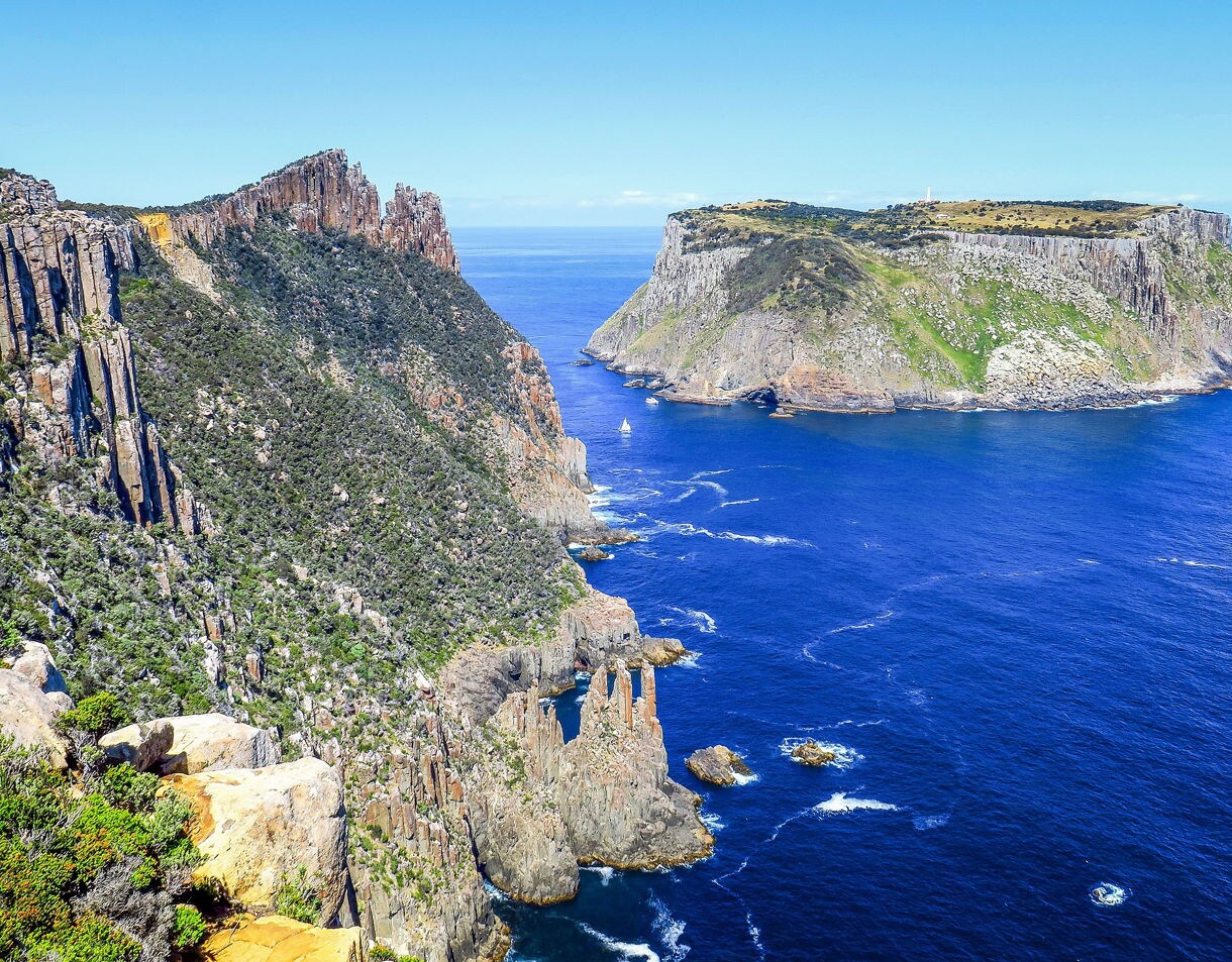 Tall column-like sea cliffs rising sharply from the ocean, with deep blue water and rugged coastline under a clear sky.