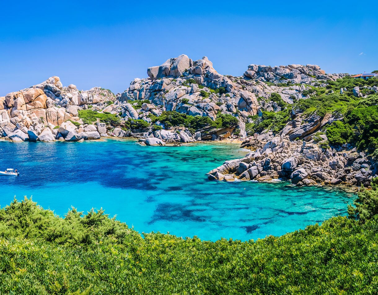 Bright turquoise cove surrounded by rocky cliffs and green shrubs under a clear blue sky.