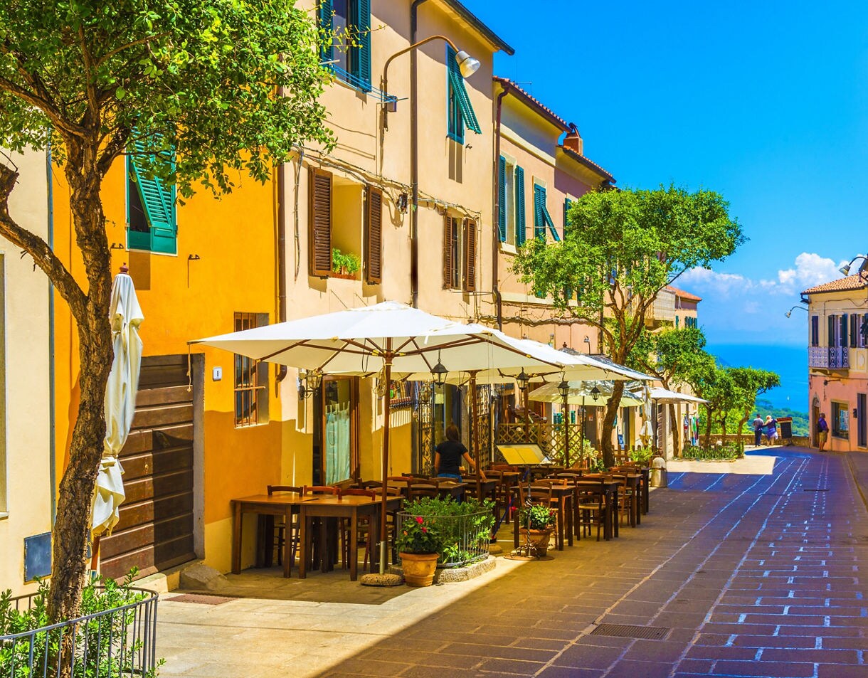 Colorful pedestrian street in Capoliveri lined with pastel buildings, outdoor café tables shaded by white umbrellas, small trees along the walkway and a view of the blue sea at the far end.