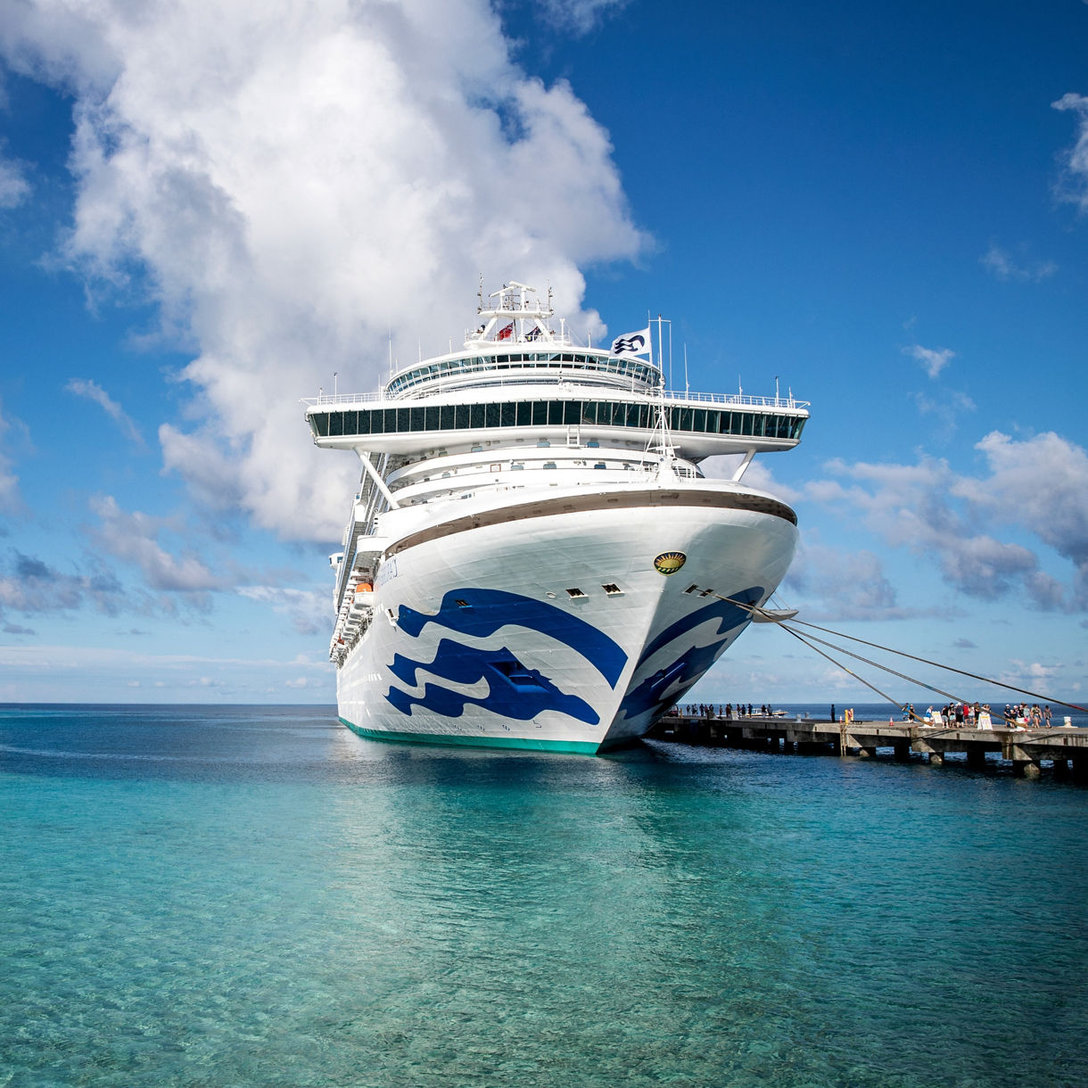 A massive white cruise ship is docked at a wooden pier in a clear tropical ocean setting. The ship features a bold blue wave graphic on its bow and is surrounded by turquoise water under a partly cloudy sky. The scene conveys a sense of leisure and travel, with the ship dominating the foreground and a distant shoreline visible. No visible numeric values or text are present.
