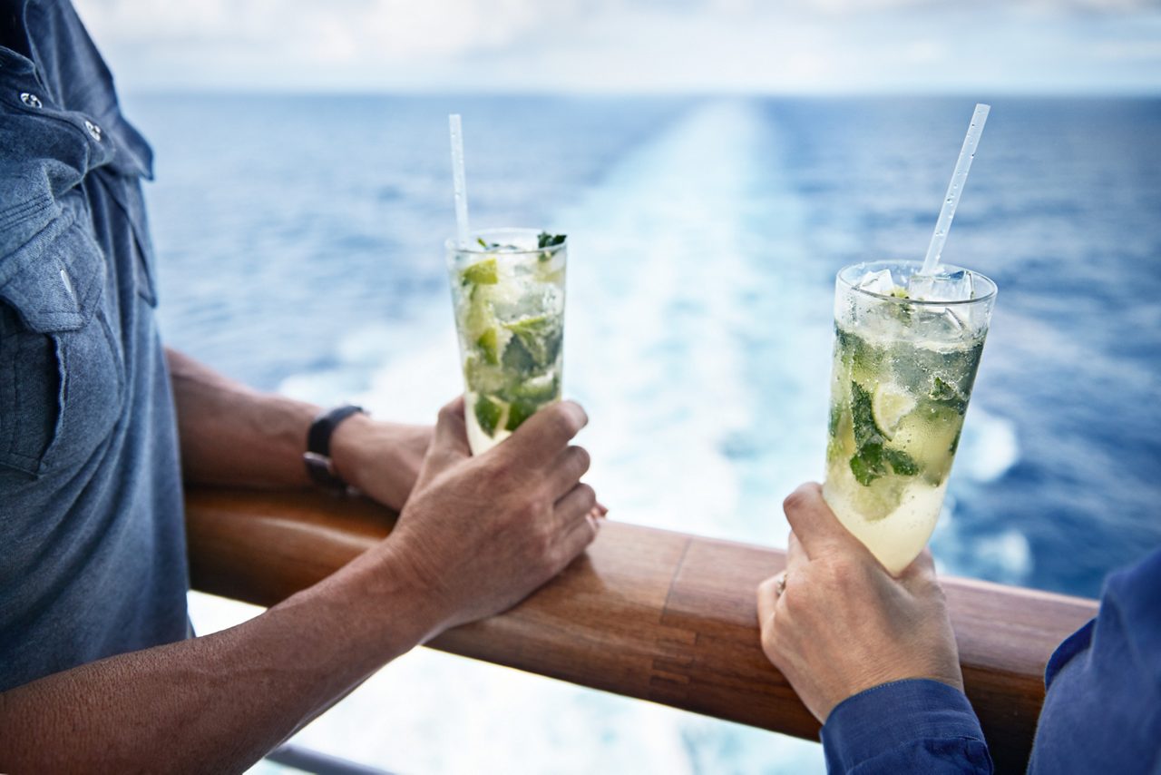 A close-up of two hands resting on a ship railing, each holding a mojito garnished with mint and a straw.