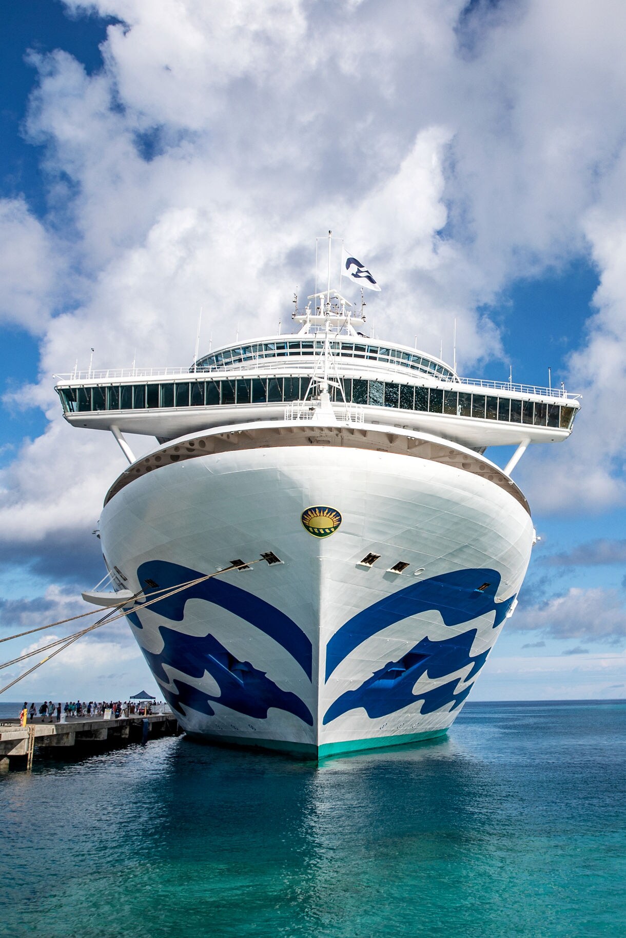 Front view of the Caribbean Princess cruise ship docked at a tropical destination, with clear turquoise waters and a partly cloudy blue sky.