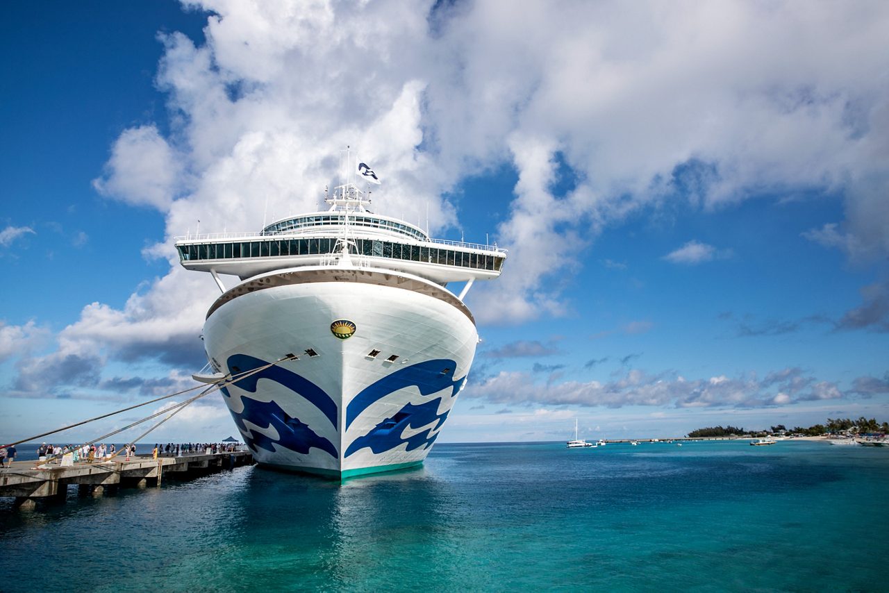 Front view of the Caribbean Princess cruise ship docked at a tropical destination, with clear turquoise waters and a partly cloudy blue sky.