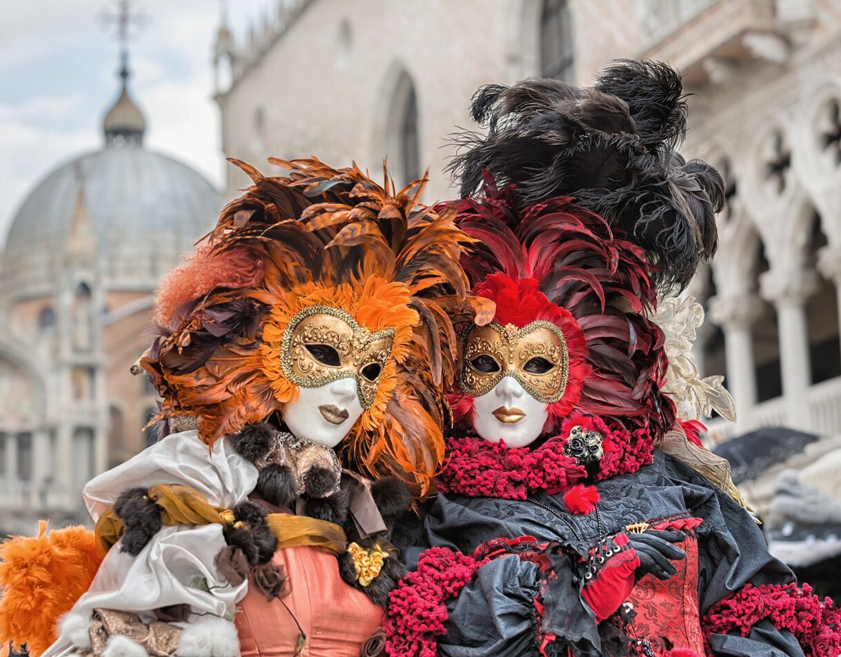 Two people wearing ornate gold masquerade masks and elaborate feathered costumes in orange, red and black, standing in front of historic Venetian architecture.