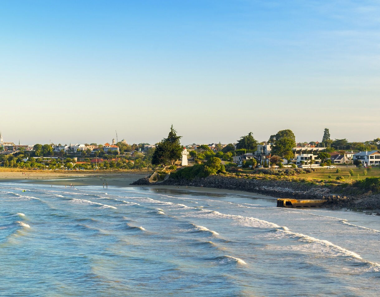 View of Caroline Bay in Timaru with soft waves, sandy beach and a line of homes and trees under clear evening light.