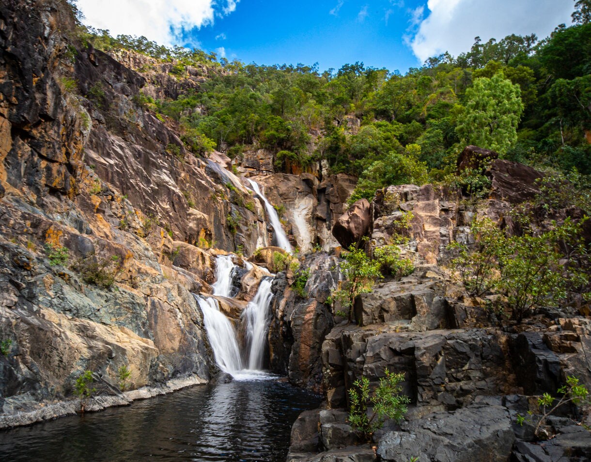 A multi-tiered waterfall flowing down rugged rock cliffs into a dark pool, surrounded by lush green trees and vegetation under a bright blue sky.