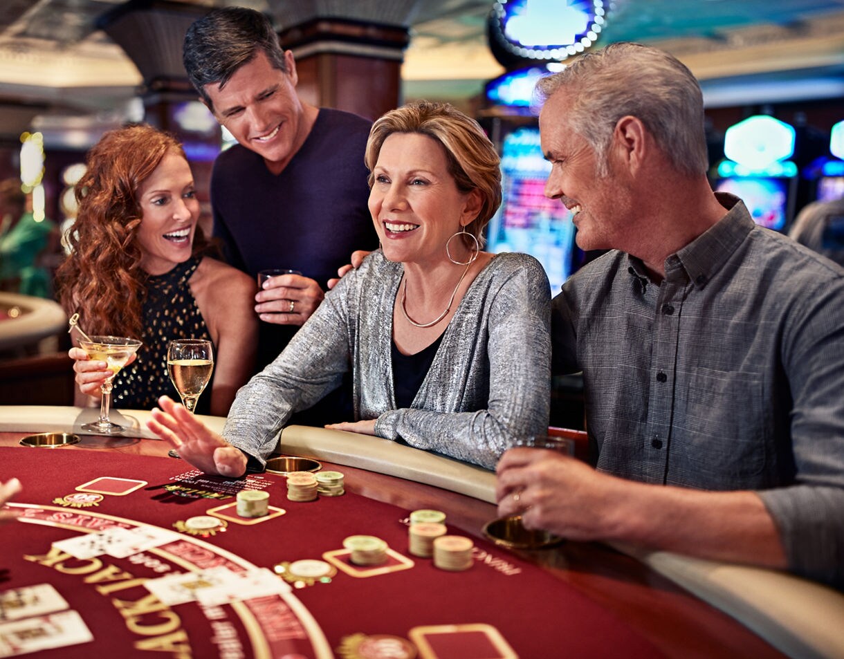  A group of four adults, two men and two women, share a laugh around a casino table on a cruise ship while enjoying drinks and gambling.