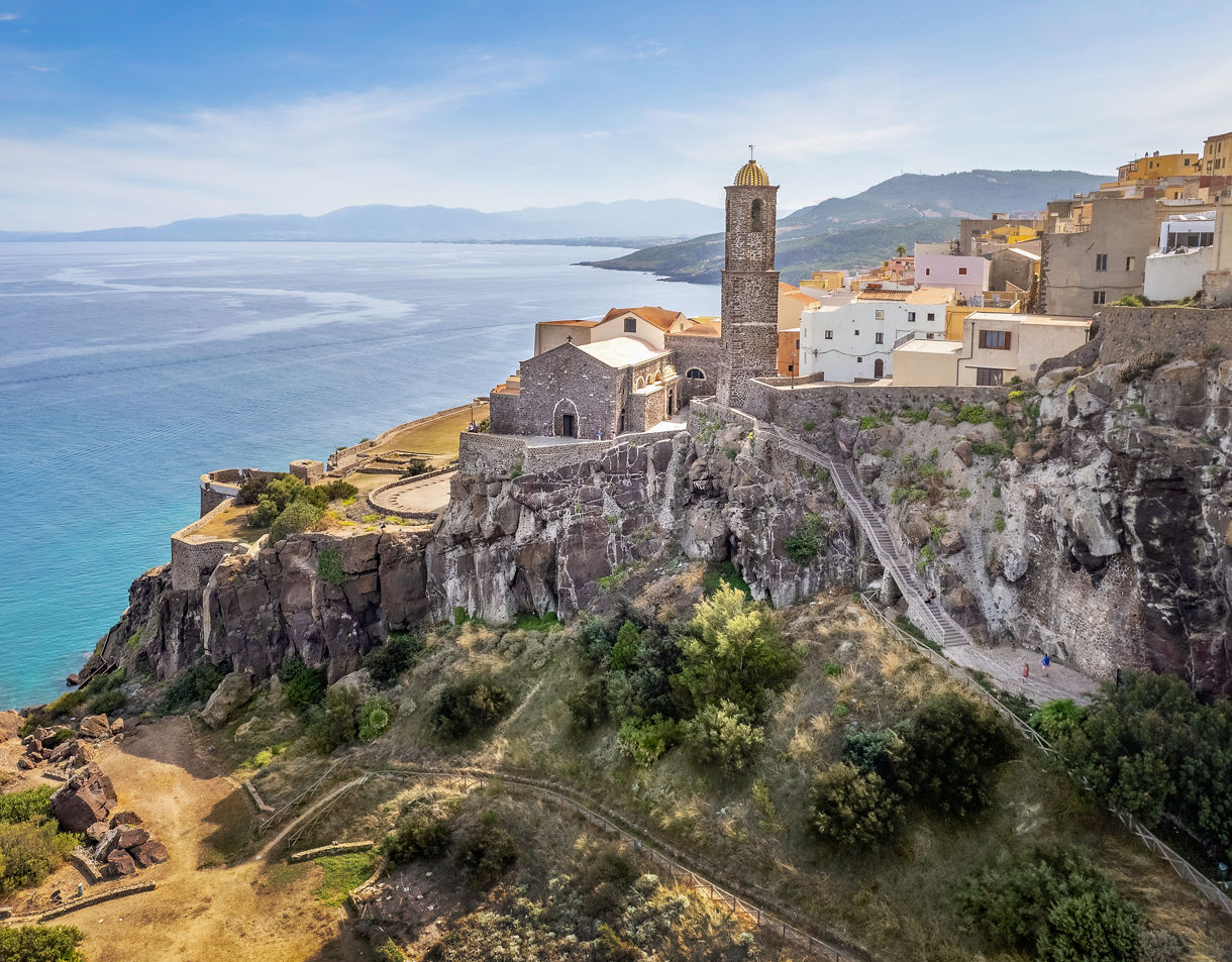 Hilltop stone buildings and a tall church tower overlooking a rocky coastline and calm blue water.